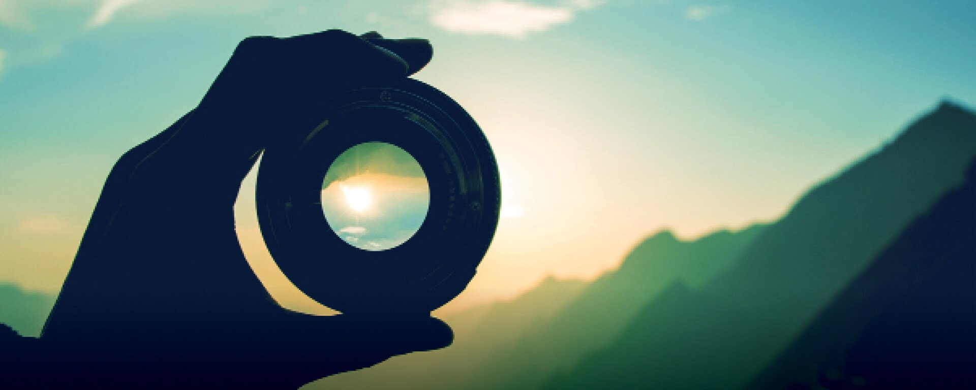 Silhouette of a hand holding a looking glass aimed at a sun setting behind mountains