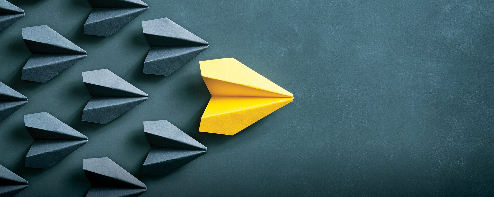 One yellow paper airplane leading a group of gray paper airplanes that blend into the gray background