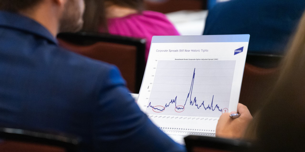 Person in blue suit holds paper with line graph showing data trends, seated among a group of people.